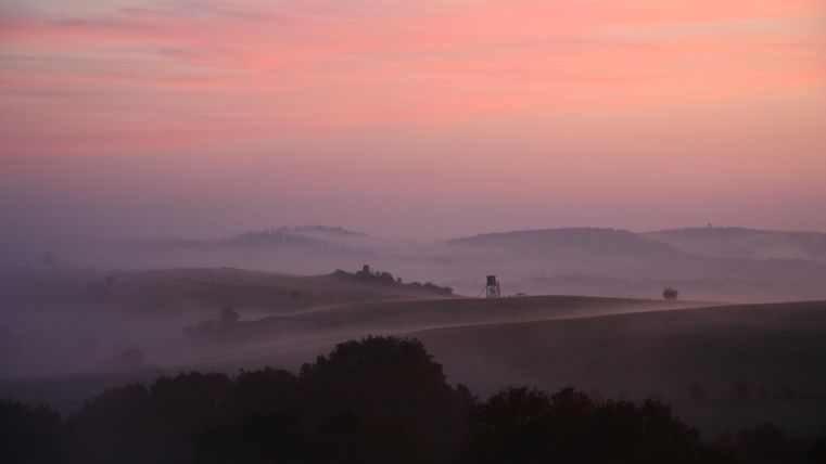 A gentle landscape with rolling hills and mist. The sky shows a beautiful play of colors at sunrise.