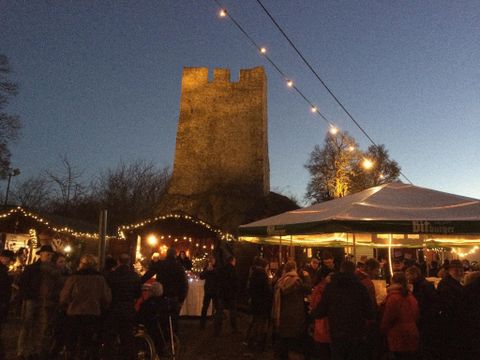 Ein festlicher Markt mit vielen Menschen vor einem historischen Turm. Die Stände sind mit Lichtern geschmückt und die Atmosphäre ist gemütlich.
