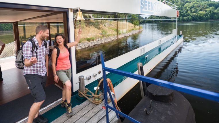 Zwei Personen steigen lachend von einem modernen Schiff mit Glaswänden aus. Eine Frau läutet eine Glocke. Im Hintergrund ist ein bewaldetes Ufer zu sehen.