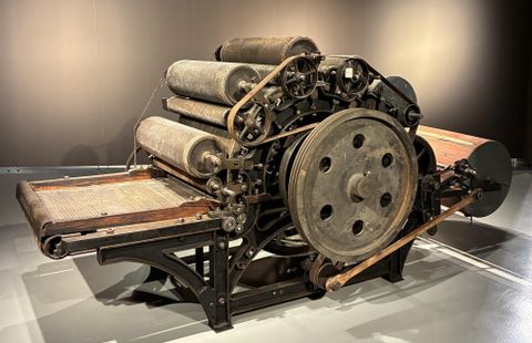 Een oude drukpers met meerdere cilinders en een robuust frame. Ze staat op een lichte vloer voor een donkere muur.