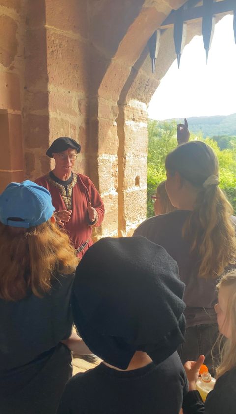 Un homme déguisé en écuyer médiéval se tient sous un arc de la Bertradaburg devant un groupe d'enfants.