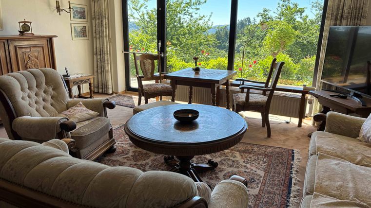 A cozy living room with comfortable furniture and a large window that offers a beautiful view of nature. In the center stands a round table as well as a dining table by the window.