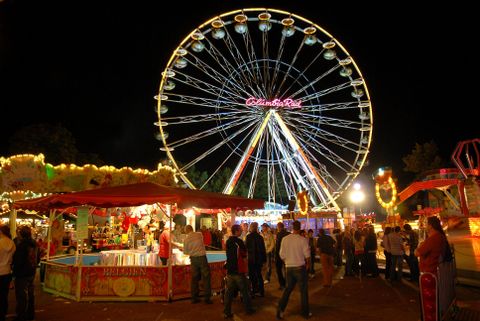 Une grande roue brille dans la nuit au-dessus d'une foire.
Les gens flânent entre les stands et les manèges.