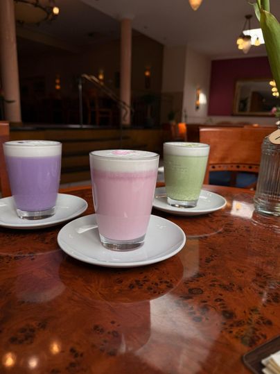 Drei Gläser mit bunten Getränken stehen auf einem Tisch. Die Farben der Getränke sind lila, pink und grün.