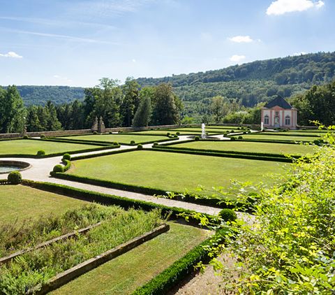 Jardin à la française avec des haies géométriques et des pelouses, au fond un petit bâtiment et des collines boisées.