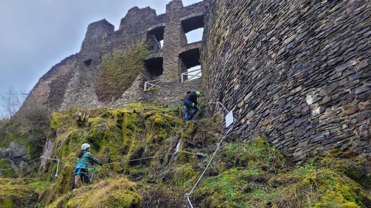 Zwei Personen klettern an einer alten Burgruine. Der Boden ist grün mit Moos und das Wetter ist bewölkt.