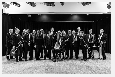 Een grote band staat samen op het podium, enkele muzikanten houden hun instrumenten vast. De groep lijkt professioneel en goed gekleed.
