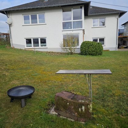 Ein Haus mit großen Fenstern steht im Hintergrund. Im Vordergrund sind ein Tisch und ein runder Grill auf dem Rasen.