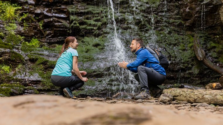 Zwei Personen hocken vor einem kleinen Wasserfall in einer natürlichen Umgebung.