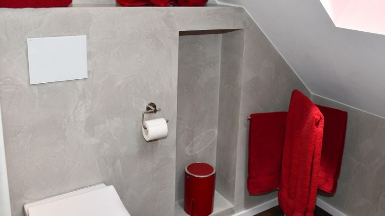A modern bathroom with a sloped ceiling and gray walls. Red towels and a red cup add colorful accents.