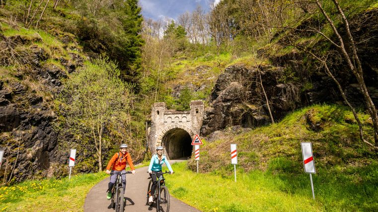 Zwei Radfahrer genießen eine malerische Landschaft auf einem Radweg. Im Hintergrund ist ein alter Tunnel und grüne Hügel zu sehen.
