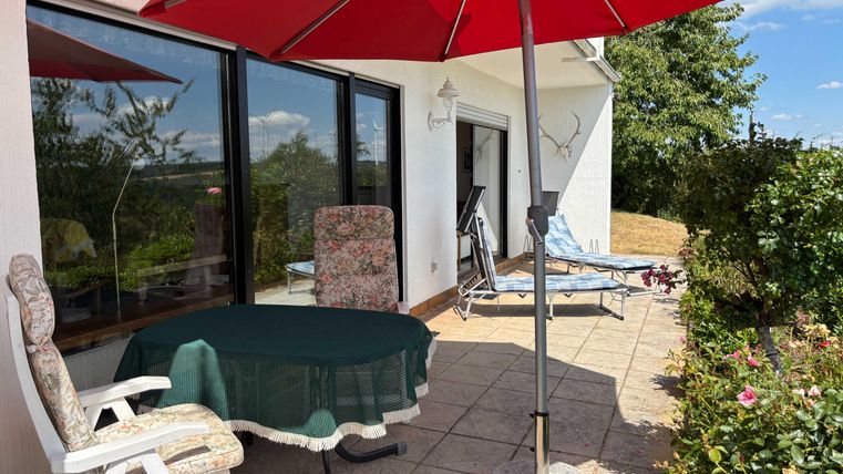 A cozy terrace with a round table and a red sun umbrella. In the background, chairs and a green garden landscape are visible.
