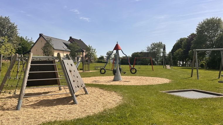 Een speeltuin met verschillende speeltoestellen zoals glijbanen en schommels. De omgeving is groen en de lucht is blauw.