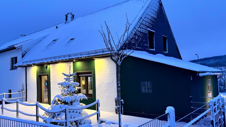 A cozy house in the snow with a illuminated entrance area. A small, decorated tree stands in front of the building.