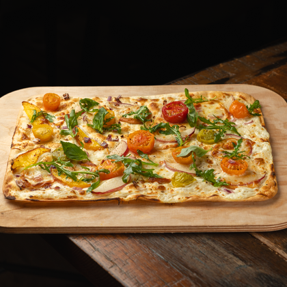 A freshly baked tarte flambée on a wooden board. It is topped with colorful regional organic tomatoes from Theo Frings and arugula.