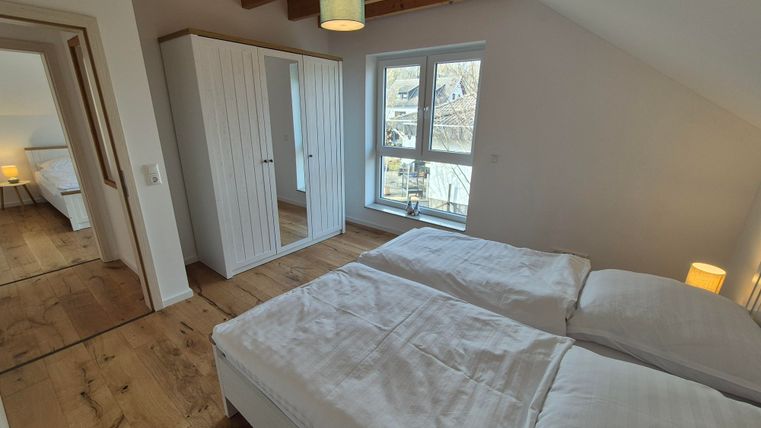 Une chambre moderne avec deux lits et une grande fenêtre. La pièce est accueillante et lumineuse, avec un sol en bois et une armoire spacieuse.