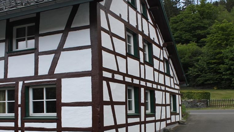 Een traditioneel vakwerkhuis met witte muren en donkere balken. Op de achtergrond zijn bomen en een rustige omgeving te zien.