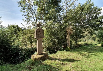 Een stenen kruis staat op een groene weide, omringd door bomen en struiken. De lucht is blauw met een paar wolken., © Tourist-Information Bitburger Land