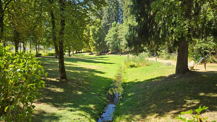 Une prairie verte entourée d'arbres, avec un petit ruisseau qui coule à travers. À l'arrière-plan, on peut voir des promeneurs.