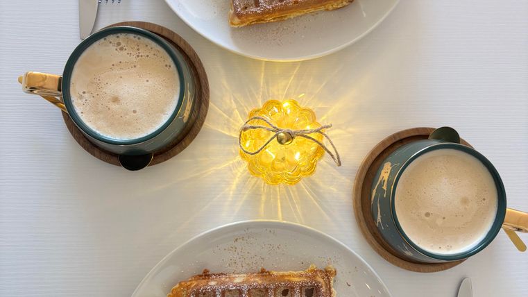 Two plates of waffles, each topped with cream and fruit, are on a table. There are also two cups of beer and a small light decoration in the middle.