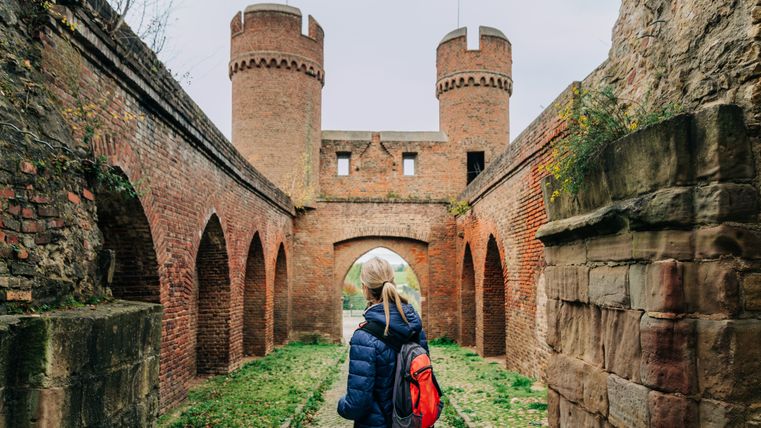 A person with a red rucksack stands in front of a historic brick archway with towers.