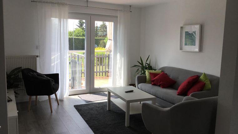 A bright living room with a gray sofa and colorful cushions. Large windows provide a view of the balcony and the greenery outside.