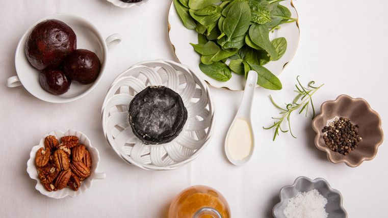 Een selectie van verse ingrediënten, waaronder spinazie, noten en Eifelfresh met pijnboomhars. Verschillende kleine schaaltjes en een glas met een oranje vloeistof zijn ook te zien.