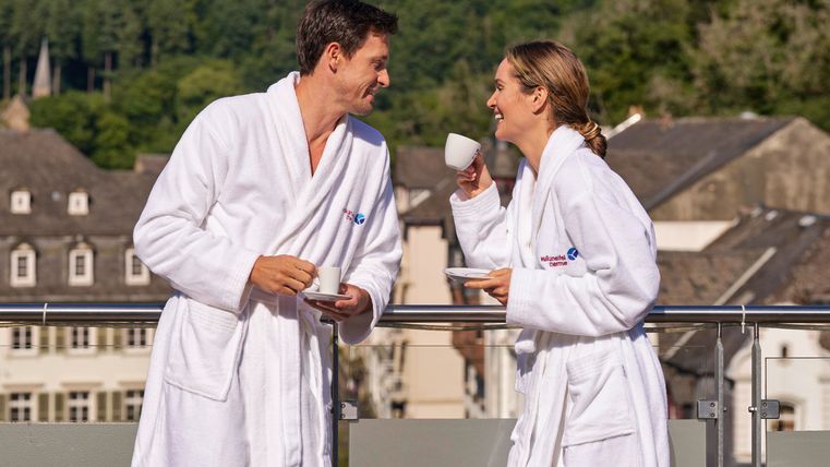 Ein lächelndes Paar in Bademänteln genießt eine Tasse Kaffee auf einem Balkon. Im Hintergrund sind malerische Gebäude und grüne Hügel sichtbar.
