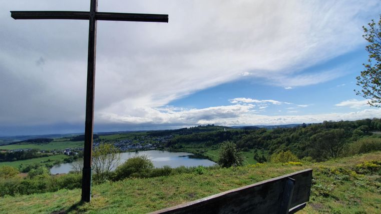 A wooden walkway leads over a calm lake, surrounded by green grass and trees. The sky is clear with a few clouds.