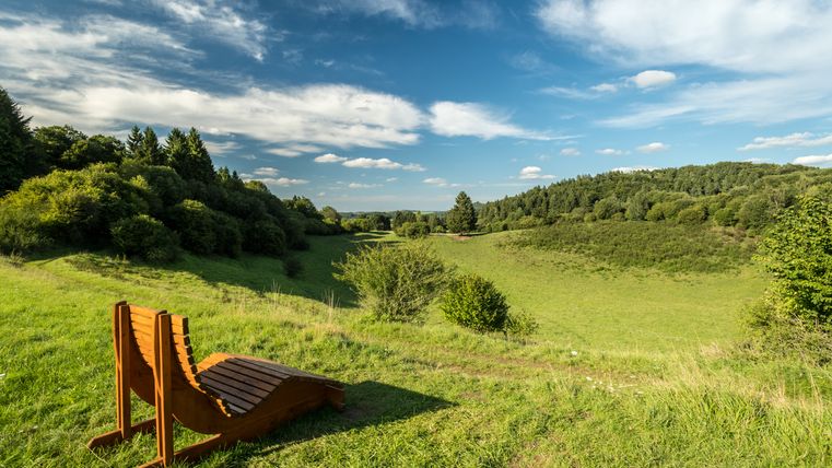 Houten ligstoel op een weiland met uitzicht op een groen landschap en blauwe lucht.