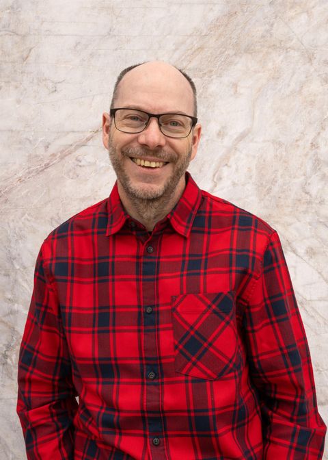 A smiling man in a plaid shirt with red and black colors stands in front of a bright, textured wall. He wears glasses and has a friendly demeanor.