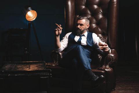 Ein stilvoller Mann sitzt in einem großen Lehnsessel und hält ein Glas Whisky in der Hand. Er trägt einen Anzug und raucht eine Zigarre, während er entspannt wirkt.