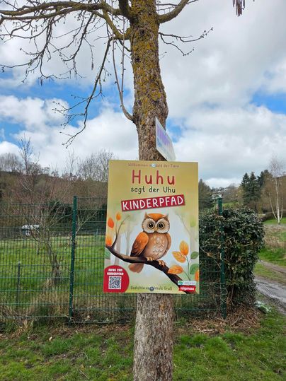 Ein Schild an einem Baum mit der Aufschrift „Huhu sagt der Uhu Kinderpfad“. Das Schild zeigt eine Illustration einer Eule mit bunten Blumen.
