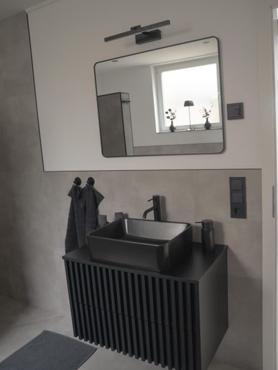 Een moderne badkamer met een elegante zwarte wastafel en een grote spiegel. De muren zijn in neutrale tinten gehouden en er zijn handdoeken en een tapijt in bijpassende kleuren.