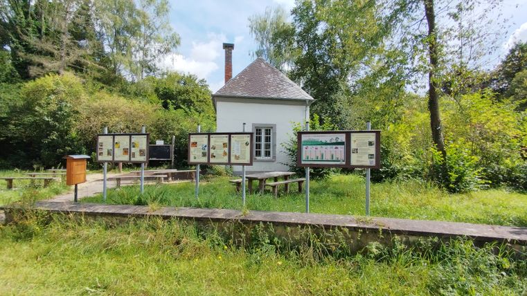 Un petit bâtiment blanc avec des panneaux d'information et des bancs devant, entouré d'arbres et d'herbe.