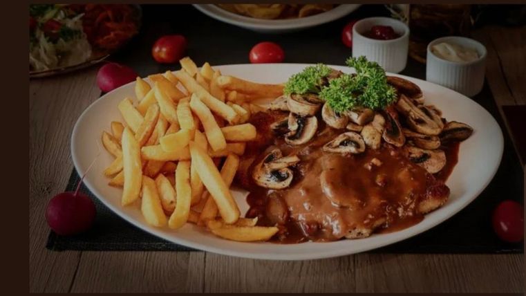 Een bord met gebraden vlees, bedekt met een saus en paddenstoelen. Daarvoor zijn er knapperige friet en verse bijgerechten.