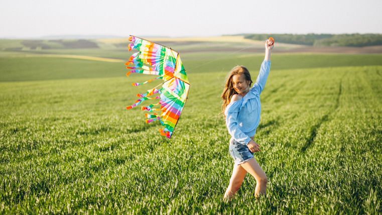 Une fille joyeuse avec un dragon coloré se tient dans un champ vert. Elle lève les bras au ciel et profite du moment.