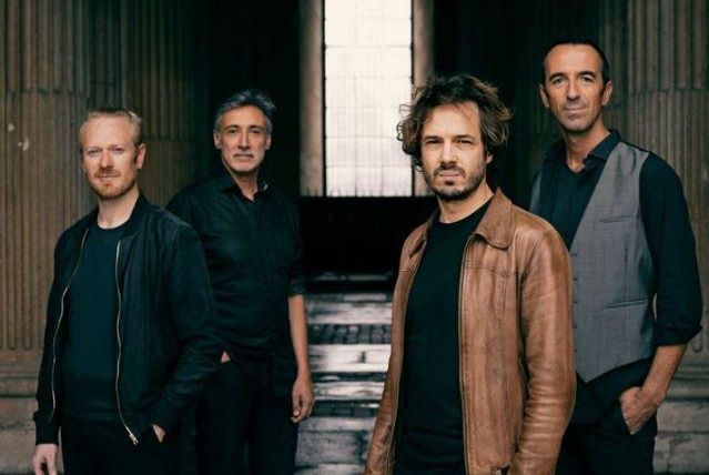 A four-member band stands in front of a window in an impressive architectural space. The members have different outfits and personal styles.