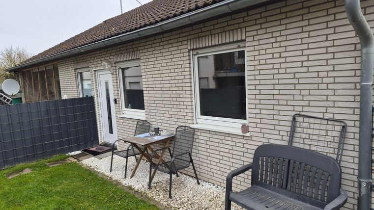 Een eenvoudig huis met een bestraatte terras en tuinmeubelen. Op de achtergrond is een grijze muur te zien.