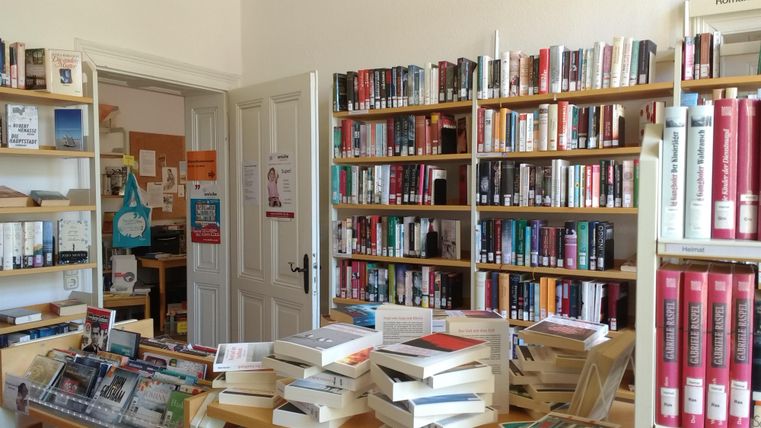 Ein gemütlicher Raum mit Bücherregalen, die voll mit verschiedenen Büchern sind. Auf einem Tisch liegen mehrere Bücher gestapelt.