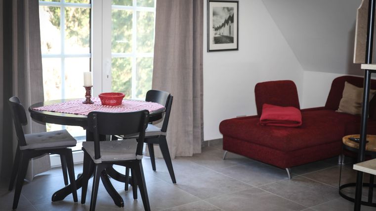 Ein gemütlicher Wohnbereich mit einem runden Tisch, Stühlen und einem roten Sofa. Große Fenster lassen viel Licht herein und schaffen eine einladende Atmosphäre.