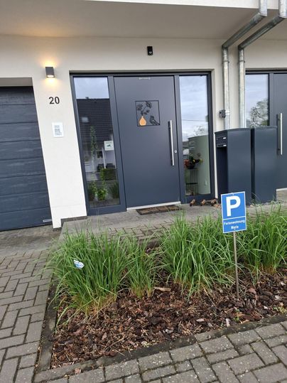 Ein modernes Haus mit einer grauen Eingangstür und einem Parkplatzschild davor. Im Vordergrund wachsen grüne Pflanzen entlang des Gehwegs.