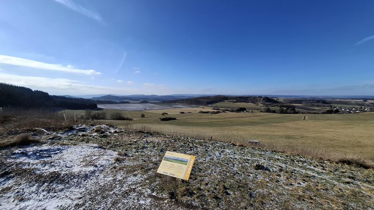 Un vaste paysage avec des collines douces et un ciel dégagé. Au premier plan se trouve un panneau d'information.