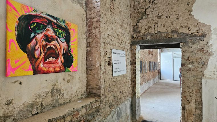 A colorful painting of a face hangs on a brick wall in an old room. Through an open passage, one can see another room in the background.