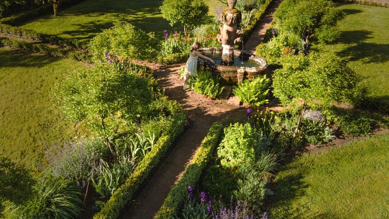Un beau jardin avec une statue au milieu et de nombreuses plantes et fleurs. Le jardin est entouré d'arbres et d'arbustes verts.