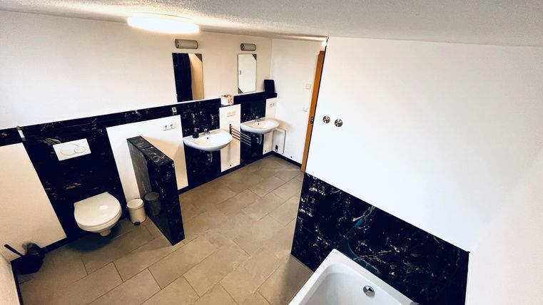 Een moderne badkamer met elegante zwarte muren en lichte tegels. Er is een bad, twee wastafels en een toilet.