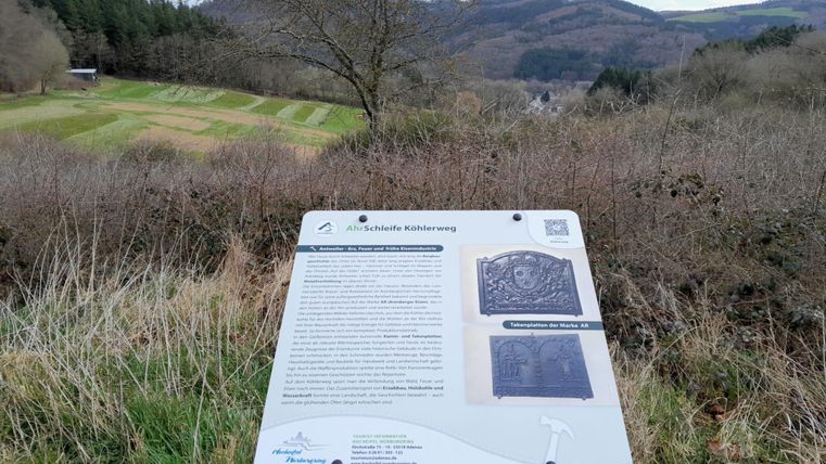 An informational sign stands in a natural landscape with gentle hills in the background. The sky is cloudy and the surroundings appear calm.
