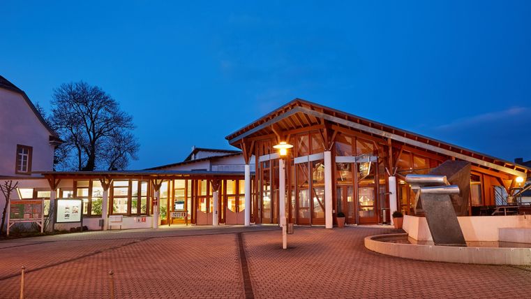 Ein modernes Gebäude bei Dämmerung mit Holz- und Glaselementen. Im Vordergrund befindet sich ein Platz mit Straßenlaternen und Bäumen.