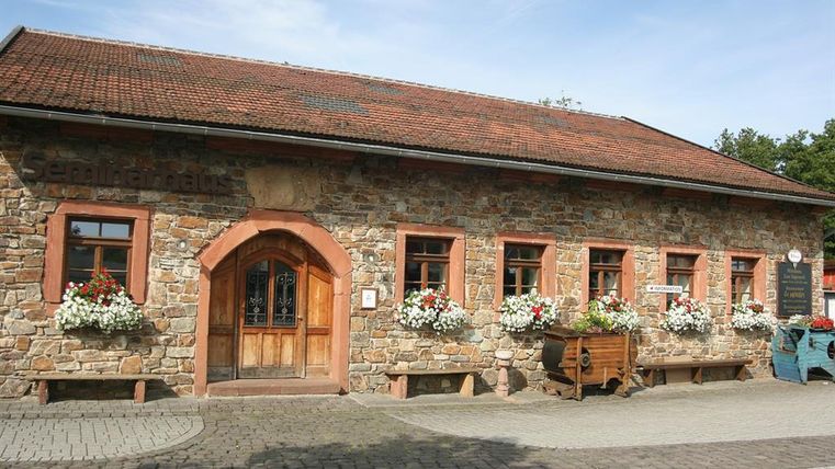 <p>Ein eingeschossiges längliches Gebäude aus Bruchsteinen und mit einem rotem Ziegeldach. Der linksgelegene Eingang hat einen Rundbogen aus rötlichem Sandstein.Die Holztür ist im oberen Bereich aus Glas und mit einem Schmiedegitter verziert. Rechts und links sind 2 feststehene Holzelemente. Links des Einganges gibt es 1 Fenster, rechts davon fünf, vierreckige Sprossenfenster, die ebenfalls von Sandsteinen umrahmt sind. Vor den Fenstern sind Blumenkästen mit weißen und roten Blumen. Der Hof vor dem Gebäude ist gepflastert.</p>