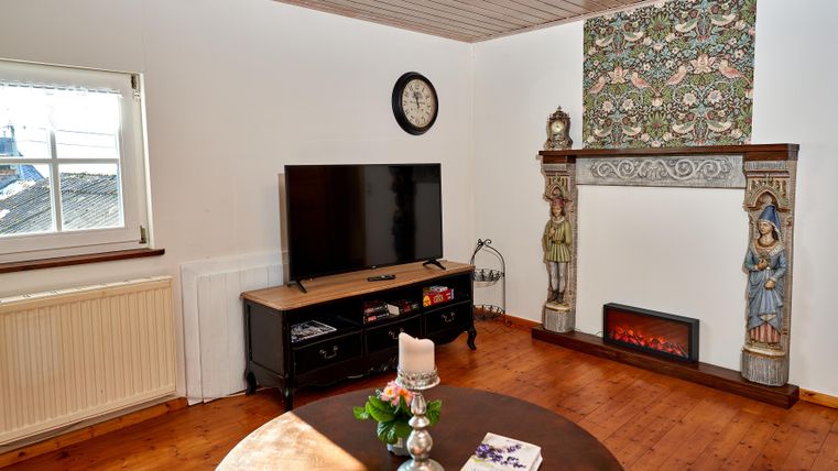 Ein gemütliches Wohnzimmer mit einem Fernseher und einem dekorativen Kamin. An der Wand befindet sich ein kunstvoll gestalteter Wandbelag und ein runder Tisch mit einer Blumenvase.